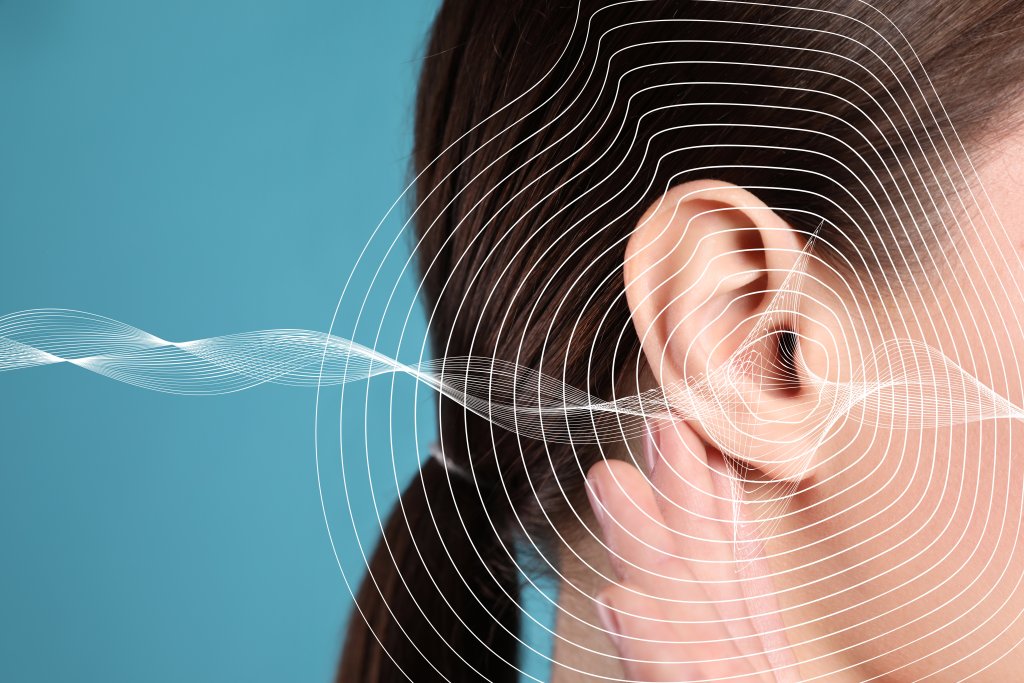 Eine Person ber&uuml;hrt ihr Ohr vor einem blauen Hintergrund, &uuml;ber dem wei&szlig;e Schallwellen abgebildet sind, die das H&ouml;ren, Tinnitus oder Nebenger&auml;usche im t&auml;glichen Leben symbolisieren.