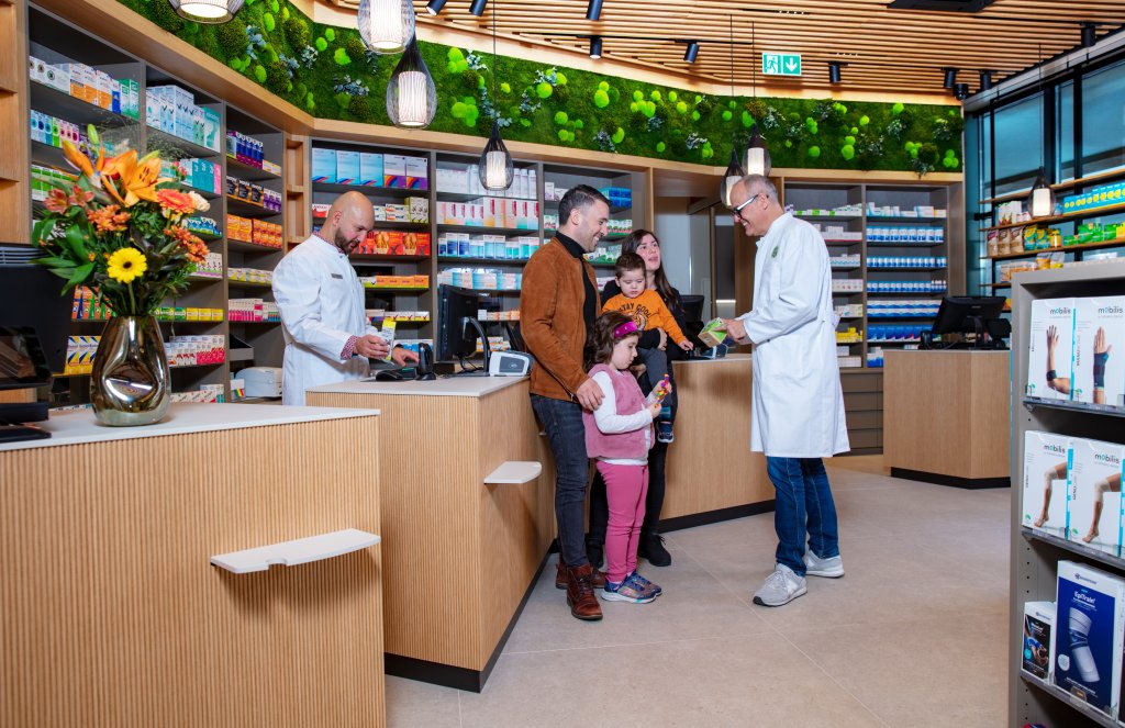 In der Apotheke Wyss unterh&auml;lt sich eine Apothekerin mit einer Familie mit zwei kleinen Kindern an der modern gestalteten Apothekentheke beim Bahnhof Baden, w&auml;hrend eine andere Apothekerin im Hintergrund eine Kundin bedient. Regale mit Medikamenten und leuchtende Blumen sind zu sehen.