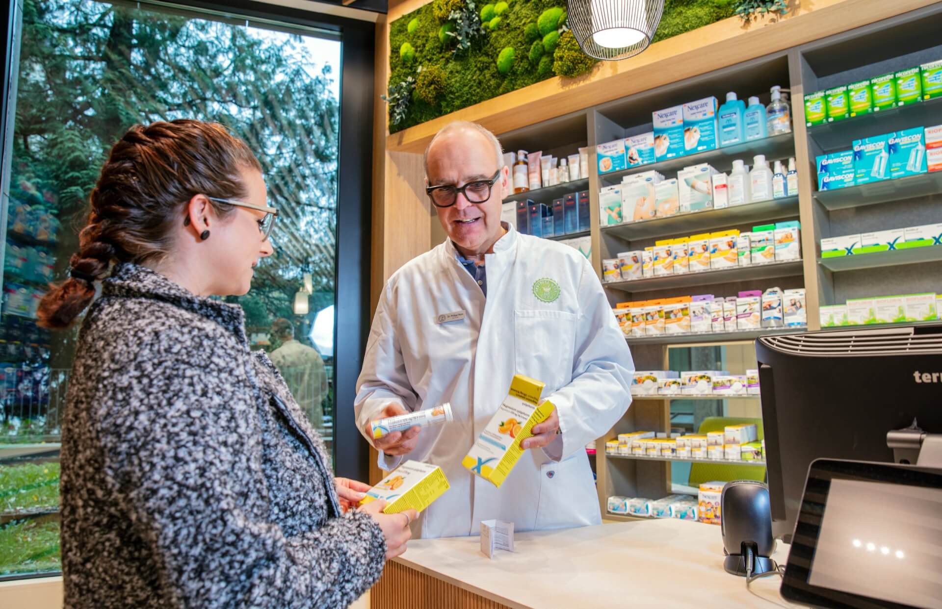 In der modernen Apotheke Wyss beim Bahnhof Baden zeigt ein Apotheker im weissen Kittel einer Frau am Schalter eine Schachtel mit Medikamenten. Dahinter schaffen Regale mit verschiedenen Medikamenten und ein Fenster mit Blick ins Gr&uuml;ne eine einladende Atmosph&auml;re.
