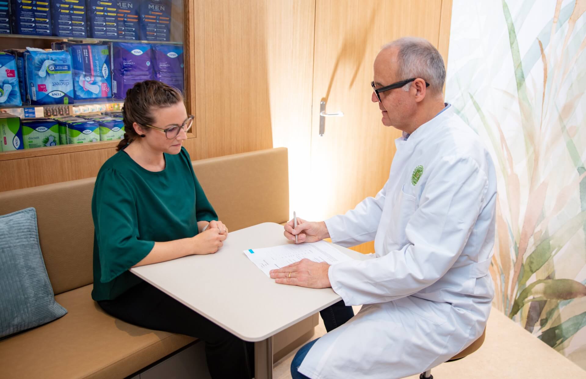 Ein Arzt im weissen Kittel sitzt an einem Tisch und schreibt in ein Formular, w&auml;hrend er sich mit einer Frau mit Brille und gr&uuml;nem Hemd in der modern gestalteten Arztpraxis der Apotheke Wyss beim Bahnhof Baden unterh&auml;lt. Im Hintergrund sind Regale mit Hygieneartikeln zu sehen.