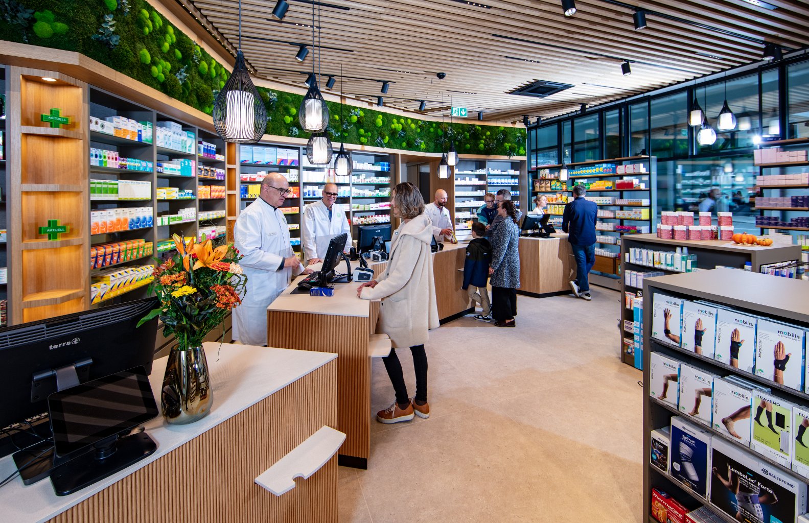 Eine modern gestaltete Apotheke Wyss nahe Bahnhof Baden, mit hellen, organisierten R&auml;umen, Holzakzenten und Pflanzen. Zwei Apotheker beraten Kunden an den Theken, w&auml;hrend weitere Besucher st&ouml;bern oder warten.