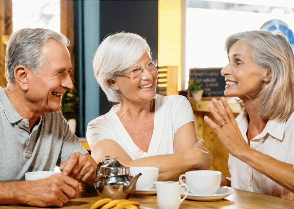 Drei ältere Erwachsene mit grauen Haaren sitzen an einem gemütlichen Cafétisch, lächeln und plaudern bei Tee und Bananen, wobei sie die Bedeutung sozialer Kontakte für die mentale Gesundheit reflektieren, während sie die Gesellschaft des anderen genießen.