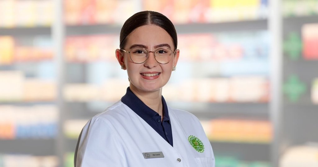 Eine junge Frau mit Brille und weißem Laborkittel steht lächelnd vor den verschwommenen Apothekenregalen. Sie hat ihr dunkles Haar zurückgesteckt und trägt ein Namensschild und einen grünen Apothekenausweis.