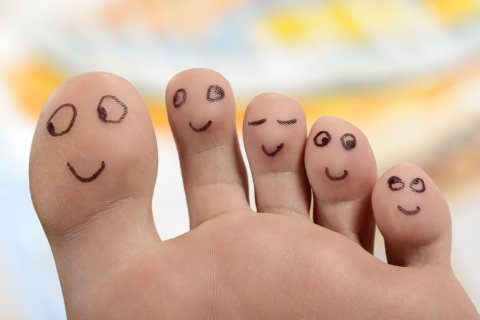 Eine Nahaufnahme von f&uuml;nf F&uuml;ssen mit fr&ouml;hlichen Gesichtern, die mit schwarzer Tinte gezeichnet sind, wobei jeder Zeh einen anderen Ausdruck zeigt. Der Hintergrund ist mit weichen Farben verschwommen, was den Eindruck erweckt, dass diese fr&ouml;hlichen Zehen in guten H&auml;nden sind.