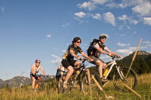 Vier sportliche Radfahrer fahren mit ihren Mountainbikes auf einem grasbewachsenen Weg unter blauem Himmel und Wolkenfetzen. Berge und B&auml;ume im Hintergrund deuten auf ein gesch&uuml;tztes Outdoor-Abenteuer hin, wobei der Sonnenschutz daf&uuml;r sorgt, dass sie auf ihrer Reise bestens gesch&uuml;tzt sind.