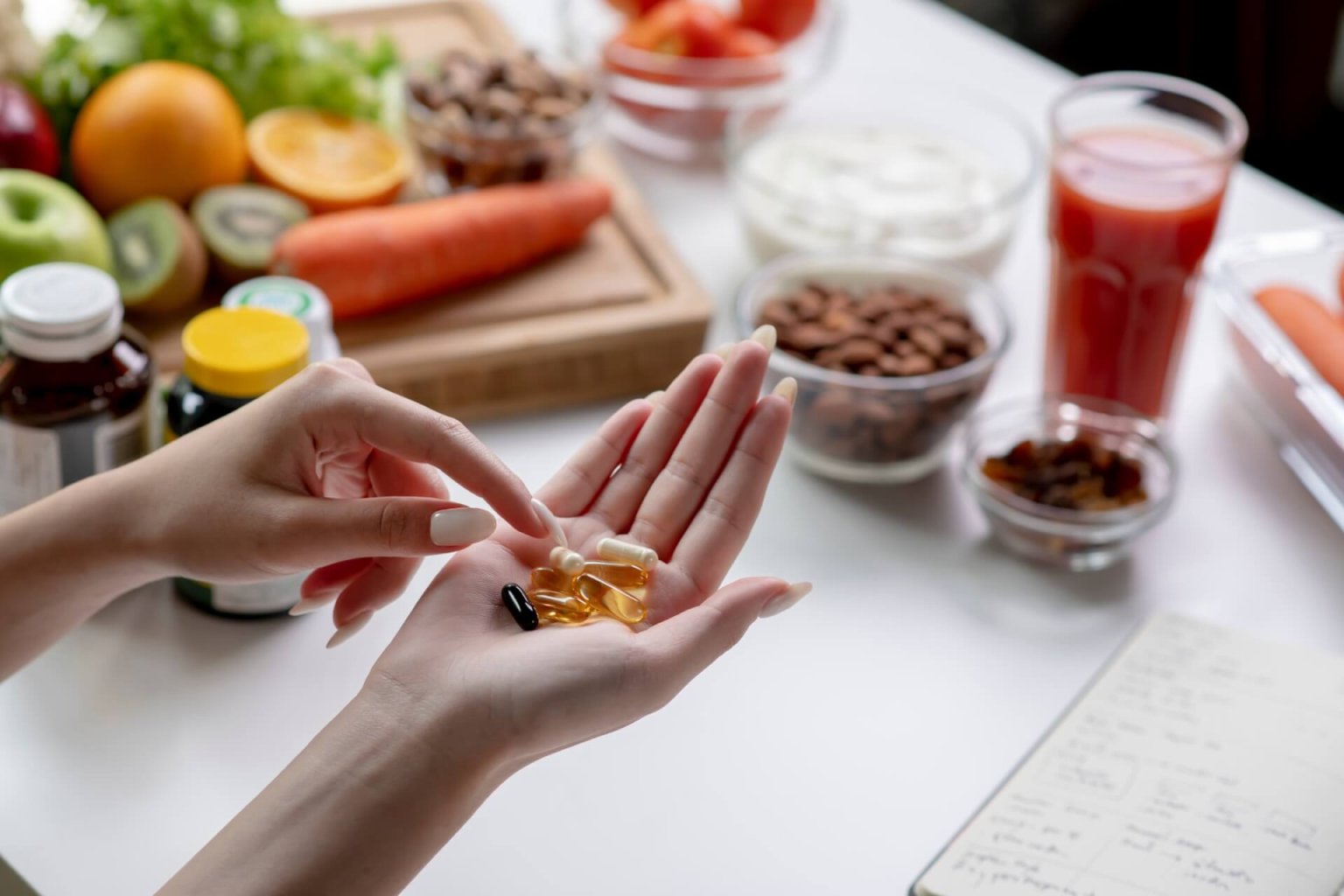 Eine Person h&auml;lt mehrere Nahrungserg&auml;nzungsmittelkapseln in einer Hand &uuml;ber einem Tisch mit Obst, Gem&uuml;se, N&uuml;ssen und einem Glas Saft, was darauf hindeutet, dass der Schwerpunkt auf Ern&auml;hrung und Gesundheit liegt.