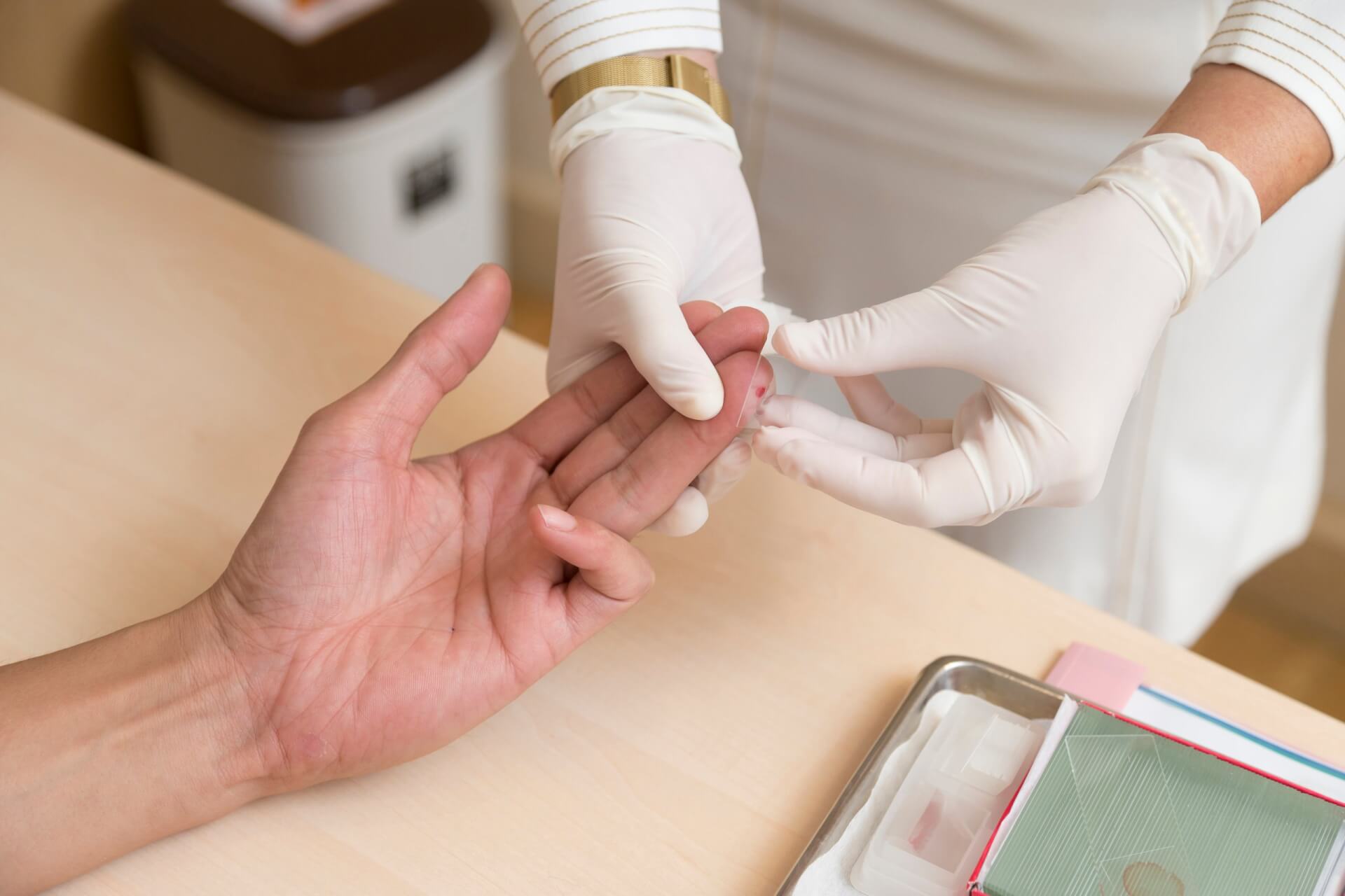 Ein Mitarbeiter des Gesundheitswesens, der Handschuhe tr&auml;gt, bereitet den Einstich in die Fingerkuppe einer Person vor, m&ouml;glicherweise f&uuml;r einen Bluttest oder eine Lipid-Messung, an einem medizinischen Untersuchungstisch mit verschiedenen Hilfsmitteln.