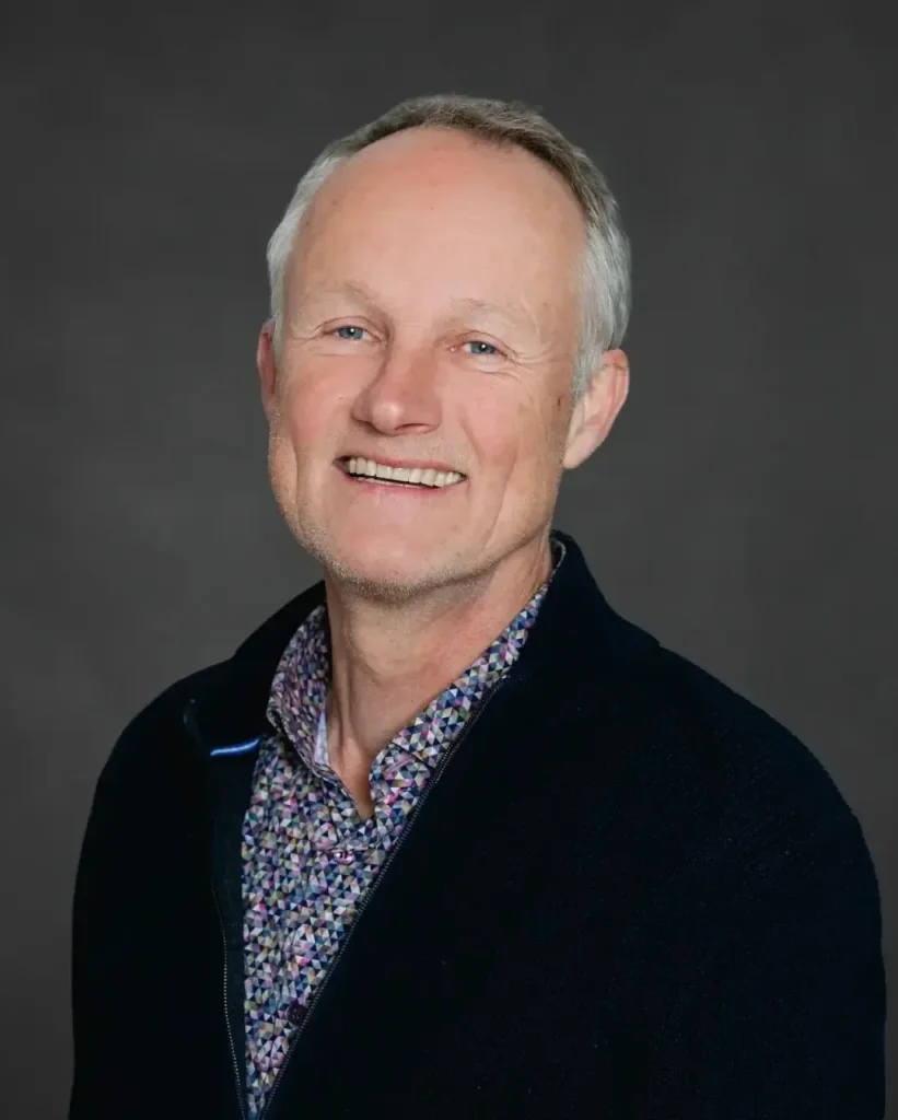 Ein Mann mittleren Alters mit kurzen grauen Haaren l&auml;chelt in die Kamera. Er tr&auml;gt eine dunkle Jacke &uuml;ber einem bunt gemusterten Hemd, das an Lobaria pulmonaria, auch bekannt als Lungenflechte, erinnert. Der Hintergrund ist einfarbig dunkelgrau.