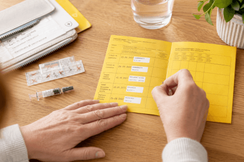 Eine Person h&auml;lt einen ge&ouml;ffneten gelben Impfausweis auf einem Holztisch neben einer Spritze, Impfstoffampullen, einem Portemonnaie, einem Glas Wasser und einer gr&uuml;nen Topfpflanze, die Impfen und Fr&uuml;hlings-M&ouml;glichkeiten in der Apotheke Wyss hervorhebt.