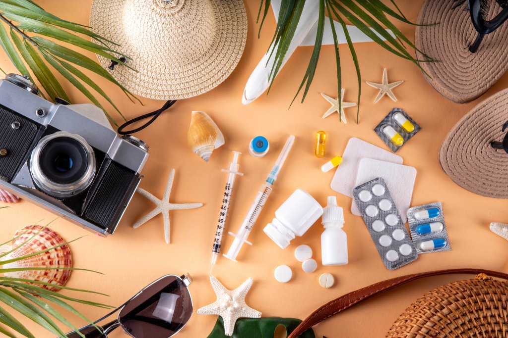 Reise- und Strandutensilien, darunter ein Fotoapparat, eine Sonnenbrille und Muscheln, sind mit Medikamenten und Pillenflaschen auf einem pfirsichfarbenen Hintergrund arrangiert, der Fr&uuml;hlings-M&ouml;glichkeiten f&uuml;r Ihre Gesundheit von der Apotheke Wyss wie Impfen und mehr hervorhebt.