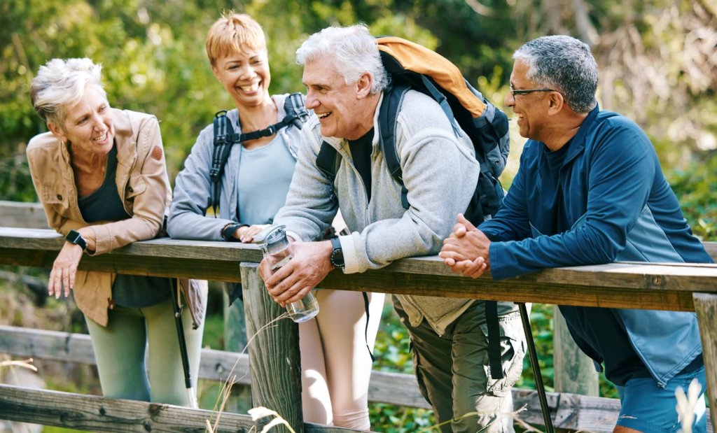 Vier ältere Erwachsene in Sportkleidung stehen auf einer Holzbrücke in einem Wald, lächeln und unterhalten sich. Einer hält eine Wasserflasche und trägt einen Rucksack. Die Gruppe scheint den Sport und die Unterstützung der Schüssler-Salze während ihrer Wanderung zu genießen.