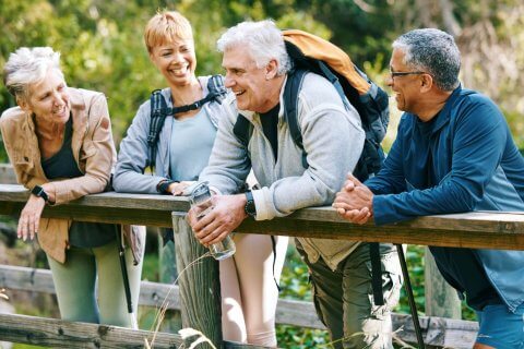 Vier &auml;ltere Erwachsene in Sportkleidung stehen auf einer Holzbr&uuml;cke in einem Wald, l&auml;cheln und unterhalten sich. Einer h&auml;lt eine Wasserflasche und tr&auml;gt einen Rucksack. Die Gruppe scheint den Sport und die Unterst&uuml;tzung der Sch&uuml;ssler-Salze w&auml;hrend ihrer Wanderung zu genie&szlig;en.