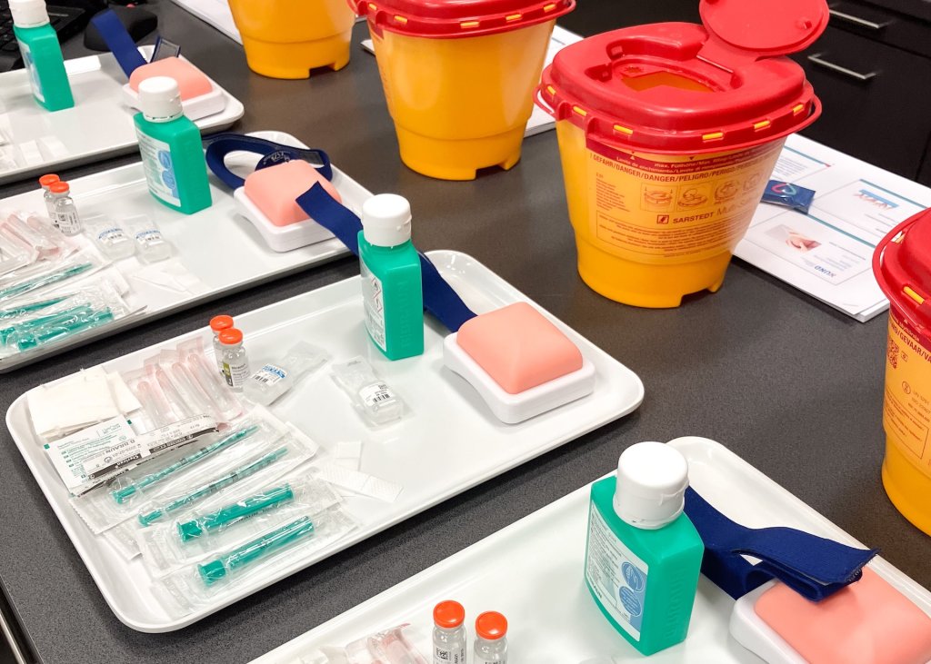 Medizinische Trainingstabletts mit Spritzen, Ampullen, antiseptischen Flaschen, &Uuml;bungspads und blauen Tourniquets sind auf einem Tisch angeordnet - ideal f&uuml;r Impfen-Demonstrationen in der Apotheke Wyss. Im Hintergrund sind auch gelbe Beh&auml;lter mit scharfen Gegenst&auml;nden und Lehrmaterial zu sehen.