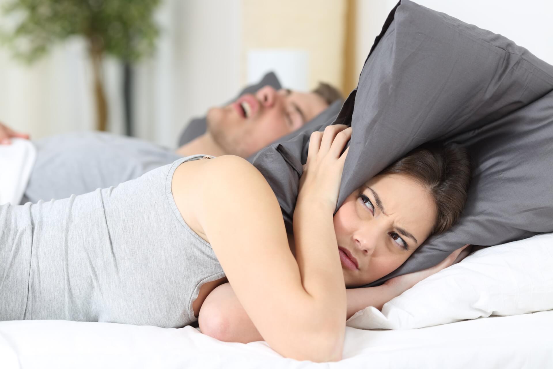Eine Frau im Bett h&auml;lt sich die Ohren mit einem Kissen zu und sieht dabei genervt aus, w&auml;hrend ein Mann neben ihr mit offenem Mund schl&auml;ft-offenbar Schnarchen auf Rezept.