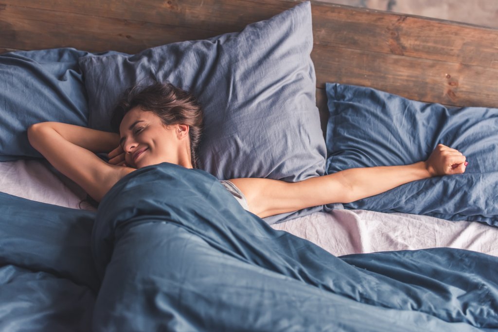 Eine Frau liegt im Bett, l&auml;chelt und streckt sich mit ausgestreckten Armen. Sie ist von einer blauen Decke zugedeckt, umgeben von blauen Kissen, und wirkt entspannt und bequem - ein Beweis daf&uuml;r, dass Schnarchen f&uuml;r erholsamen Schlaf m&ouml;glich ist.