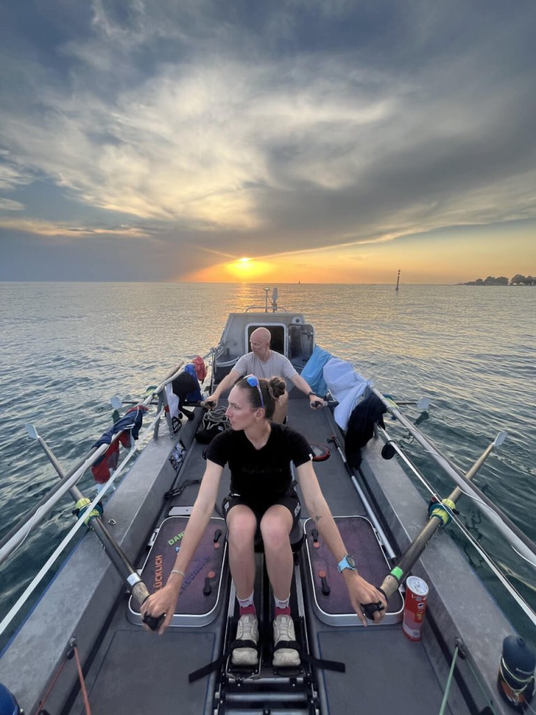 Zwei Menschen rudern bei Sonnenuntergang auf ruhigem Wasser in einem Boot. Der Himmel bietet mit seinen Wolken und dem Sonnenlicht ein dramatisches Bild. UNITED4HOPE spiegelt sich in ihrer stillen St&auml;rke wider &ndash; eine Person blickt nach vorne und h&auml;lt die Ruder fest, w&auml;hrend die andere hinten sitzt und nach unten schaut. Die Ruderausr&uuml;stung und die Kleidung liegen im Boot und symbolisieren Entschlossenheit und Zusammenhalt auf ihrer Reise.