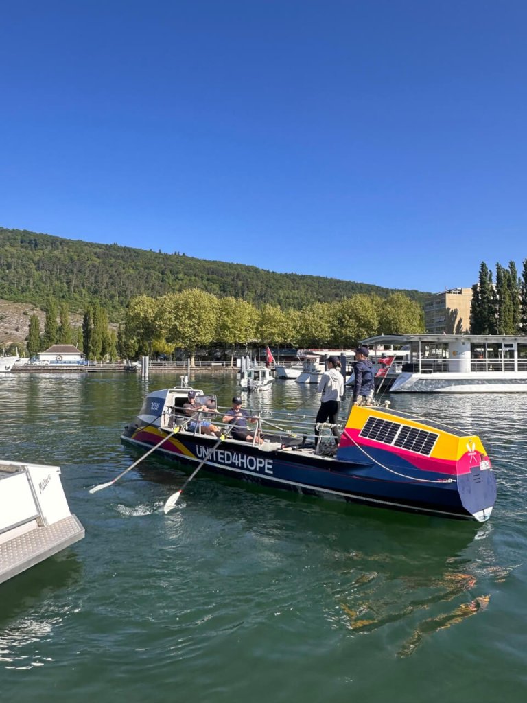 Eine Gruppe rudert mit ihrem Boot &bdquo;UNITED4HOPE&ldquo; &uuml;ber einen sonnigen See, vorbei an Anlegestellen und wei&szlig;en Booten, vor dem Hintergrund gr&uuml;ner H&uuml;gel und B&auml;ume unter einem strahlend blauen Himmel.