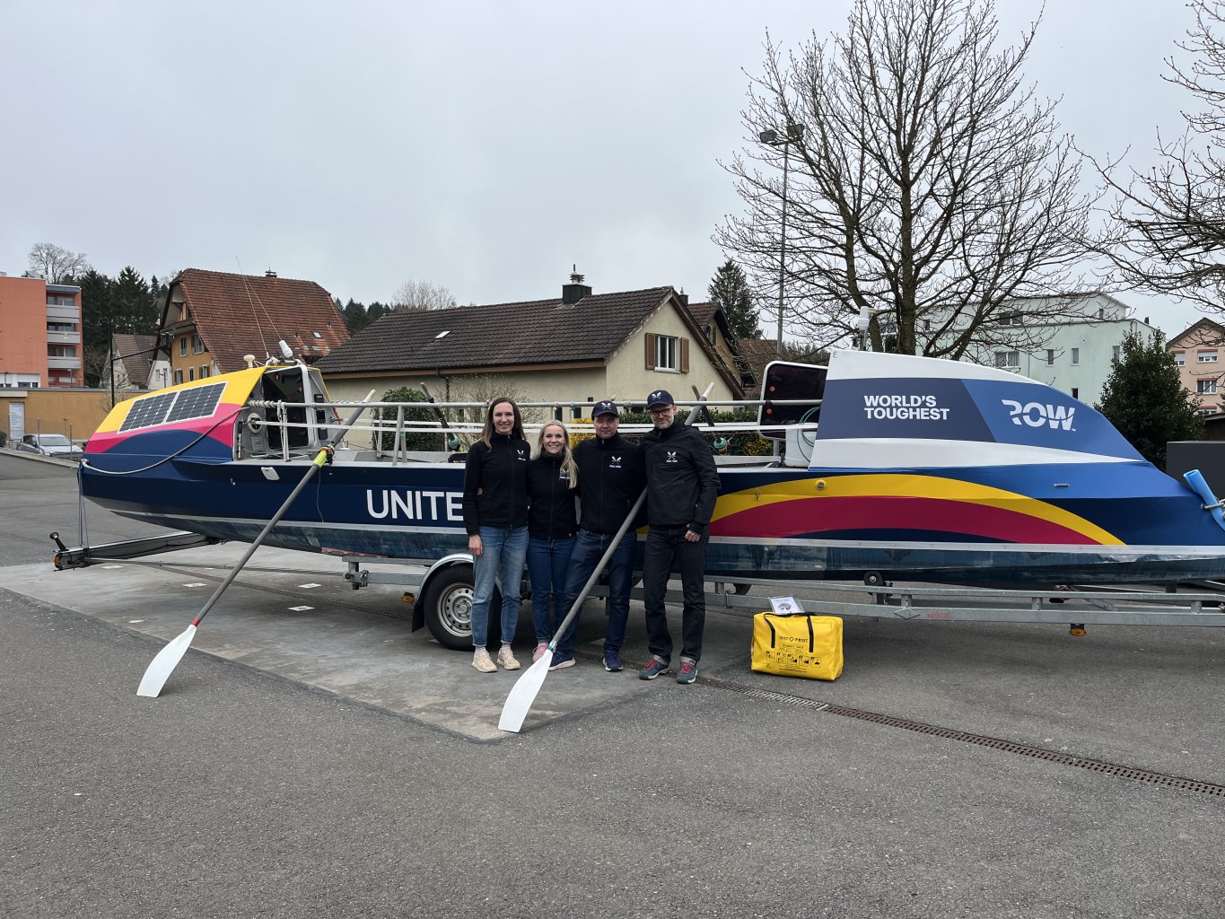 Vier Menschen stehen vor einem bunten Ruderboot auf einem Anh&auml;nger, auf dem stolz der Schriftzug &bdquo;WORLDS TOUGHEST ROW&ldquo; prangt. Die Gruppe vertritt UNITED4HOPE und bereitet sich auf eine inspirierende Reise vor. Im Hintergrund sind H&auml;user und kahlen B&auml;ume zu sehen, w&auml;hrend vorne zwei wei&szlig;e Ruder auf dem Boden liegen.