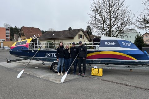 Vier Menschen stehen vor einem bunten Ruderboot auf einem Anh&auml;nger, auf dem stolz der Schriftzug &bdquo;WORLDS TOUGHEST ROW&ldquo; prangt. Die Gruppe vertritt UNITED4HOPE und bereitet sich auf eine inspirierende Reise vor. Im Hintergrund sind H&auml;user und kahlen B&auml;ume zu sehen, w&auml;hrend vorne zwei wei&szlig;e Ruder auf dem Boden liegen.