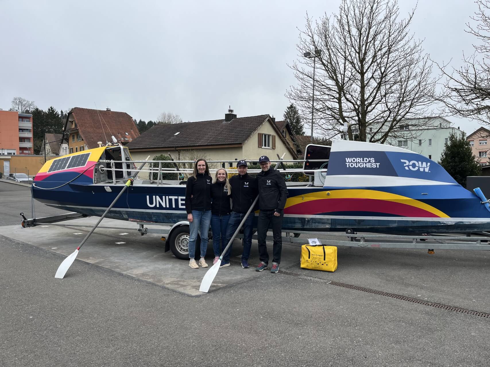 Vier Menschen stehen vor einem bunten Ruderboot auf einem Anh&auml;nger, auf dem stolz der Schriftzug &bdquo;WORLDS TOUGHEST ROW&ldquo; prangt. Die Gruppe vertritt UNITED4HOPE und bereitet sich auf eine inspirierende Reise vor. Im Hintergrund sind H&auml;user und kahlen B&auml;ume zu sehen, w&auml;hrend vorne zwei wei&szlig;e Ruder auf dem Boden liegen.